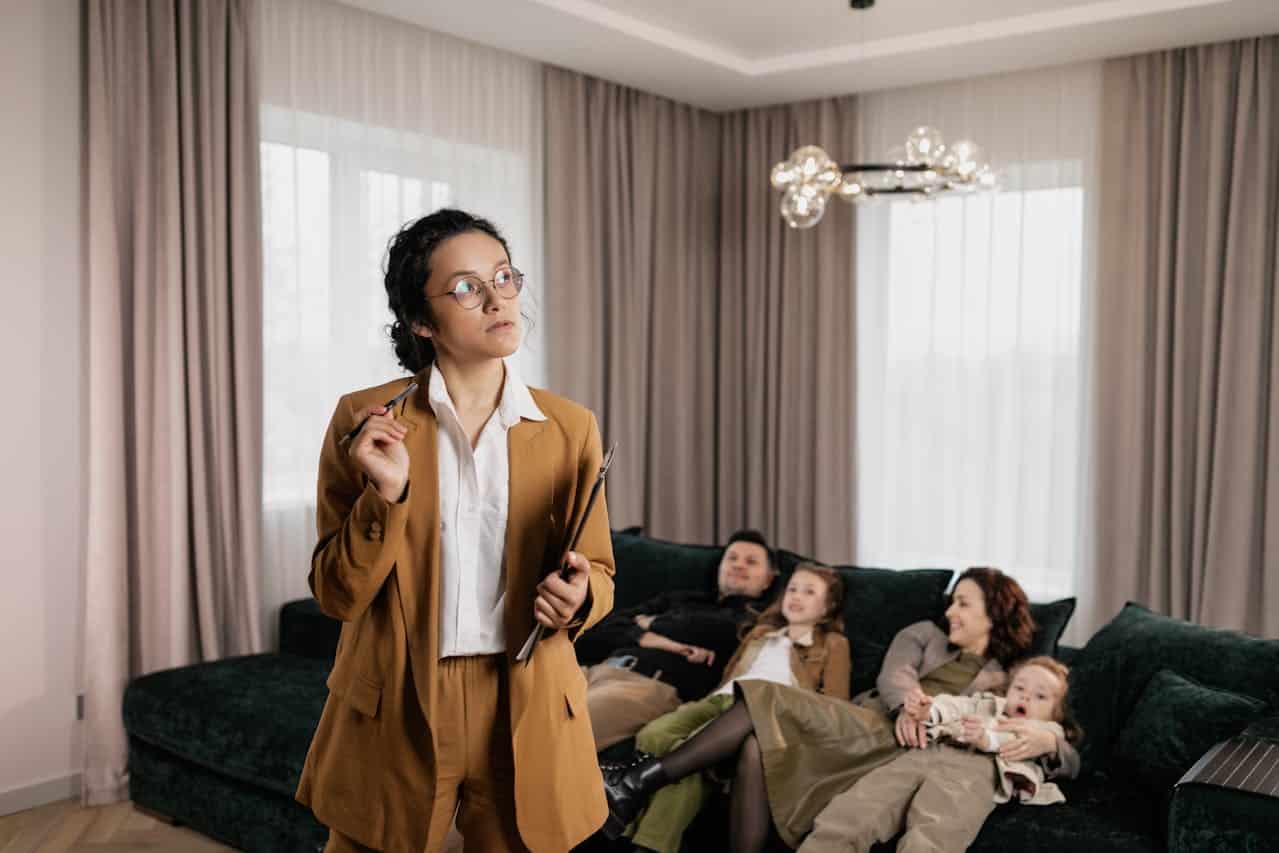 Woman in business attire holding a clipboard and pen, contemplating in a modern living room, with a family relaxing on a green sofa, illustrating personalized real estate transactions and Contract for Deed agreements.