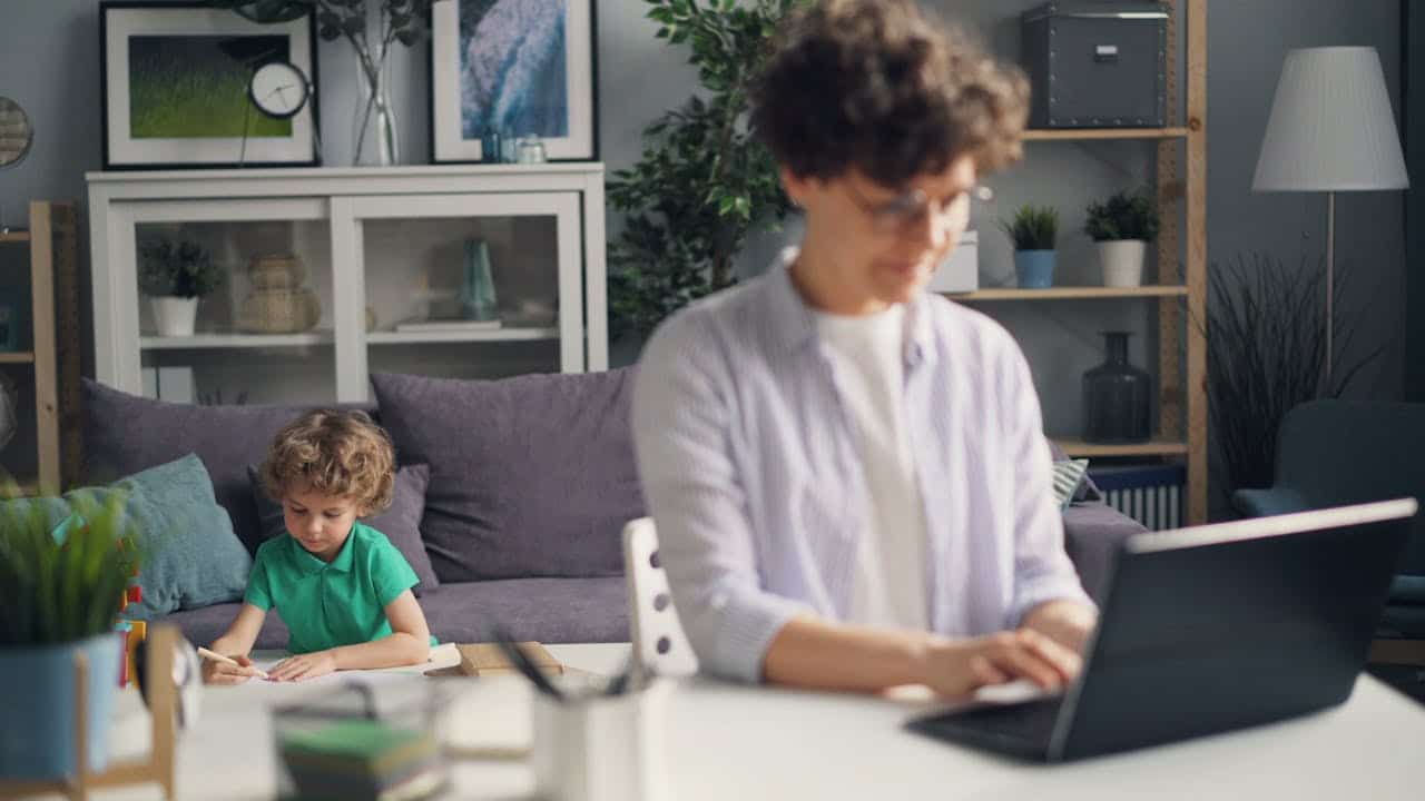 Woman working on a laptop in a home office while a child draws at a nearby table, illustrating a family-friendly remote work environment.