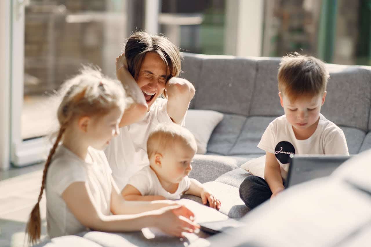 Family enjoying time together at home, with a mother playfully covering her ears while sitting on a couch with three children, two of whom are engaged with electronic devices, illustrating a lively home environment.