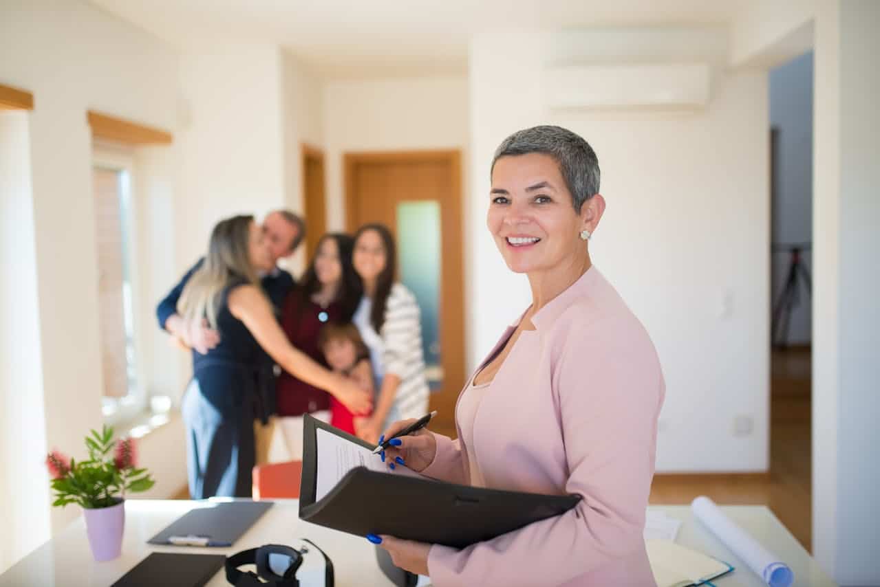 Realtor smiling while holding a contract in a bright room, with a family celebrating in the background, illustrating the process of a Contract for Deed transaction.