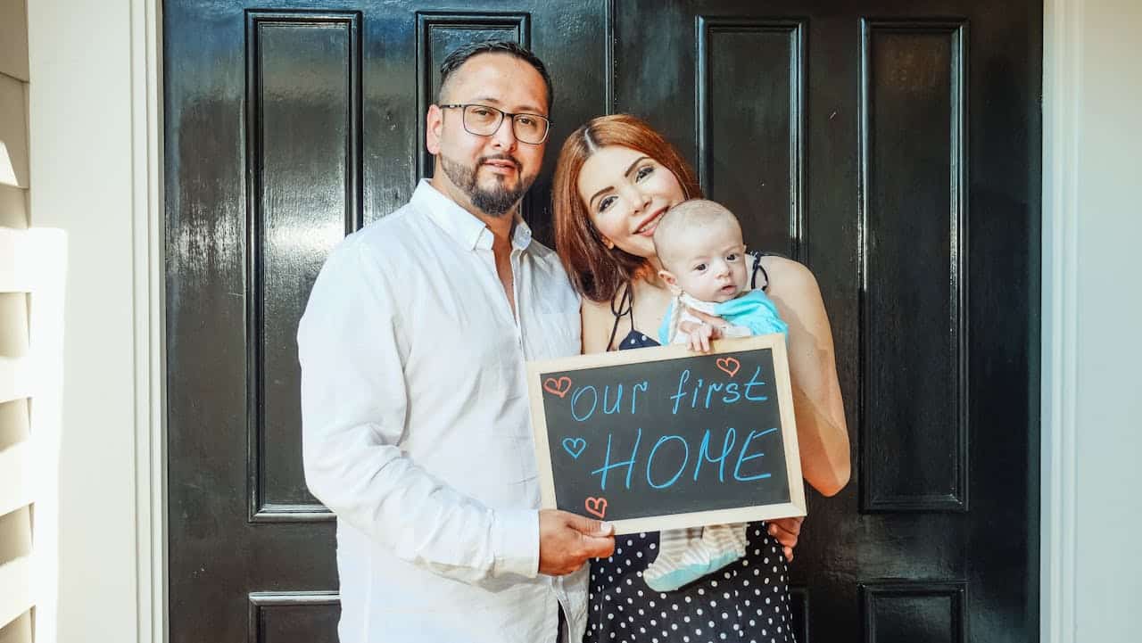 Family celebrating homeownership, holding a sign that says "our first HOME" in front of a black door, representing the success of a Contract for Deed arrangement.