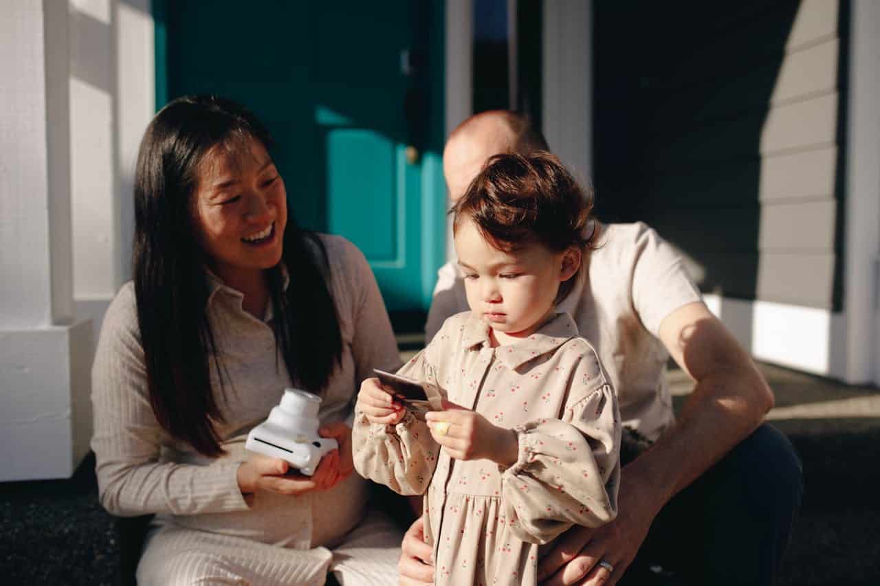 Family enjoying time together outdoors, woman smiling while holding a camera, child examining a photo, with a man seated beside them.