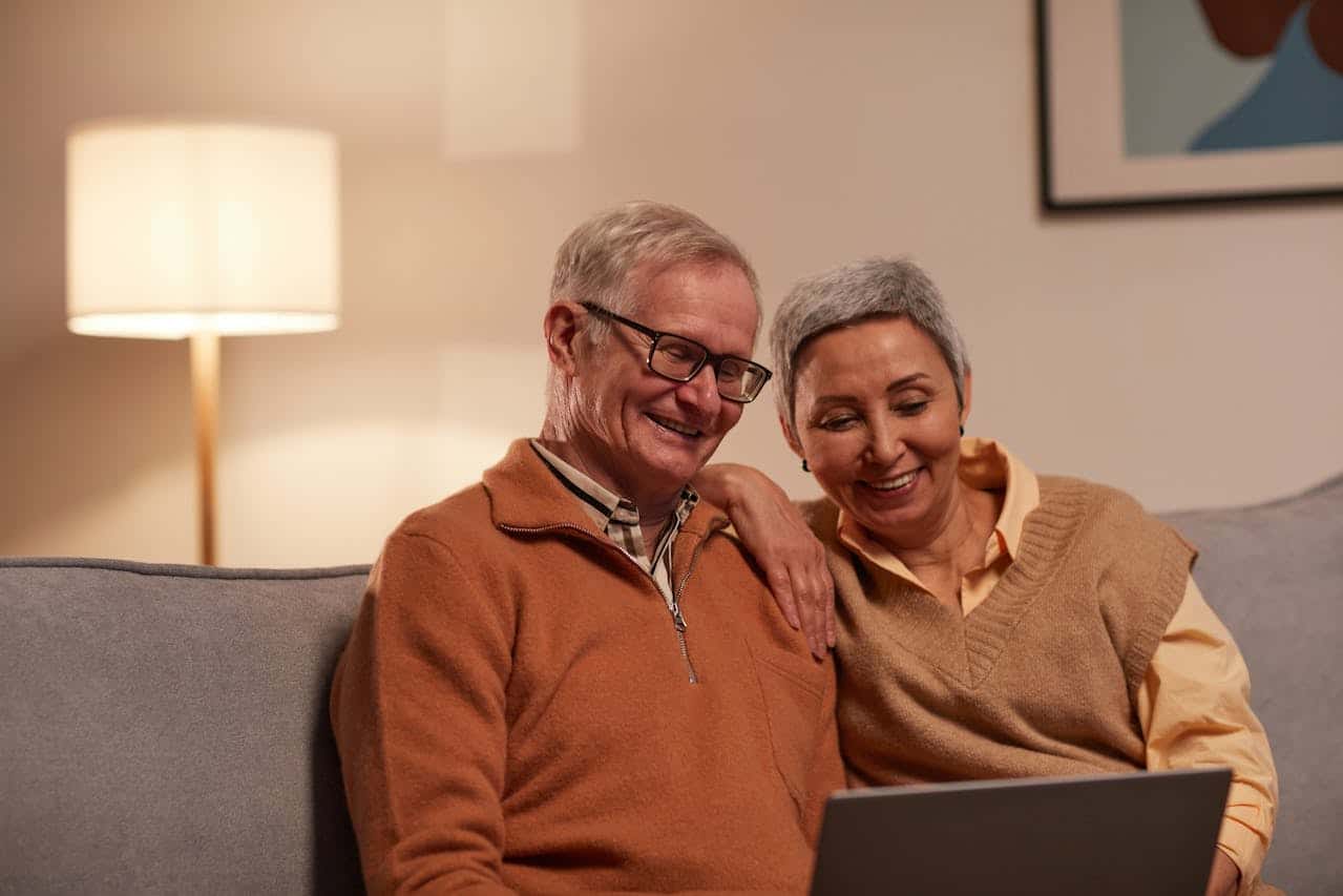 Elderly couple smiling and reviewing information on a laptop in a cozy living room setting, highlighting the importance of guidance in real estate transactions like Contract For Deed agreements.