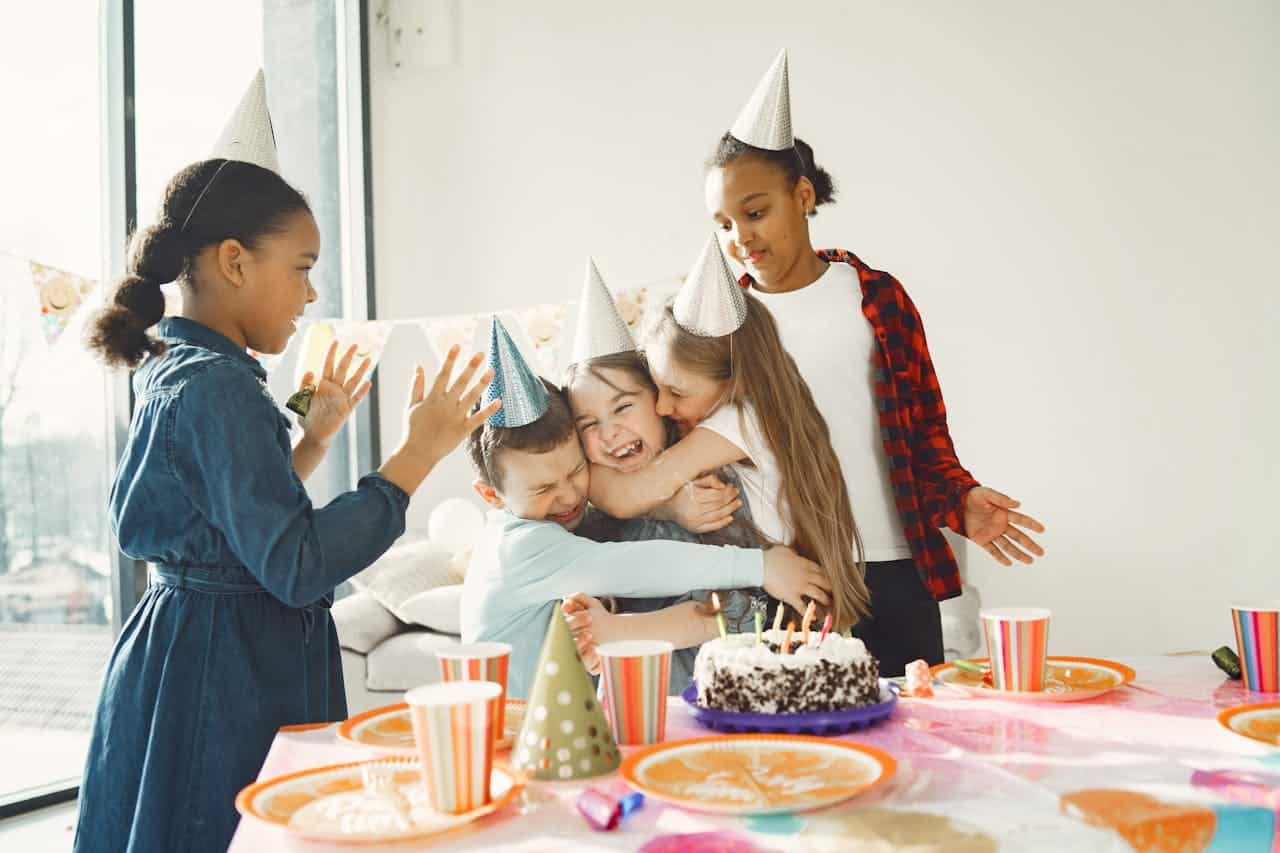 Children celebrating a birthday party, wearing party hats, hugging around a cake, with colorful decorations and snacks on a table.