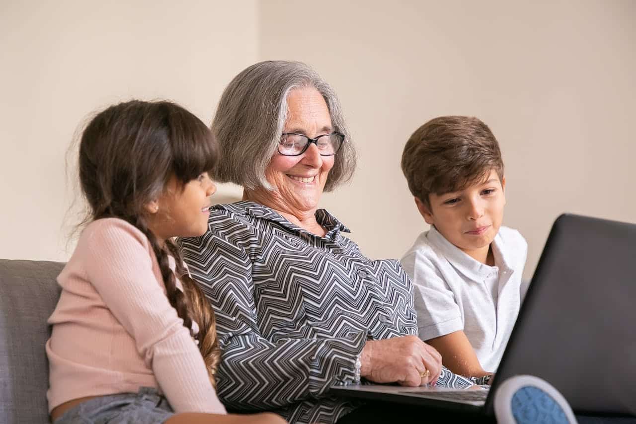Grandmother smiling while using a laptop with two children, engaging in a shared moment of learning or entertainment.