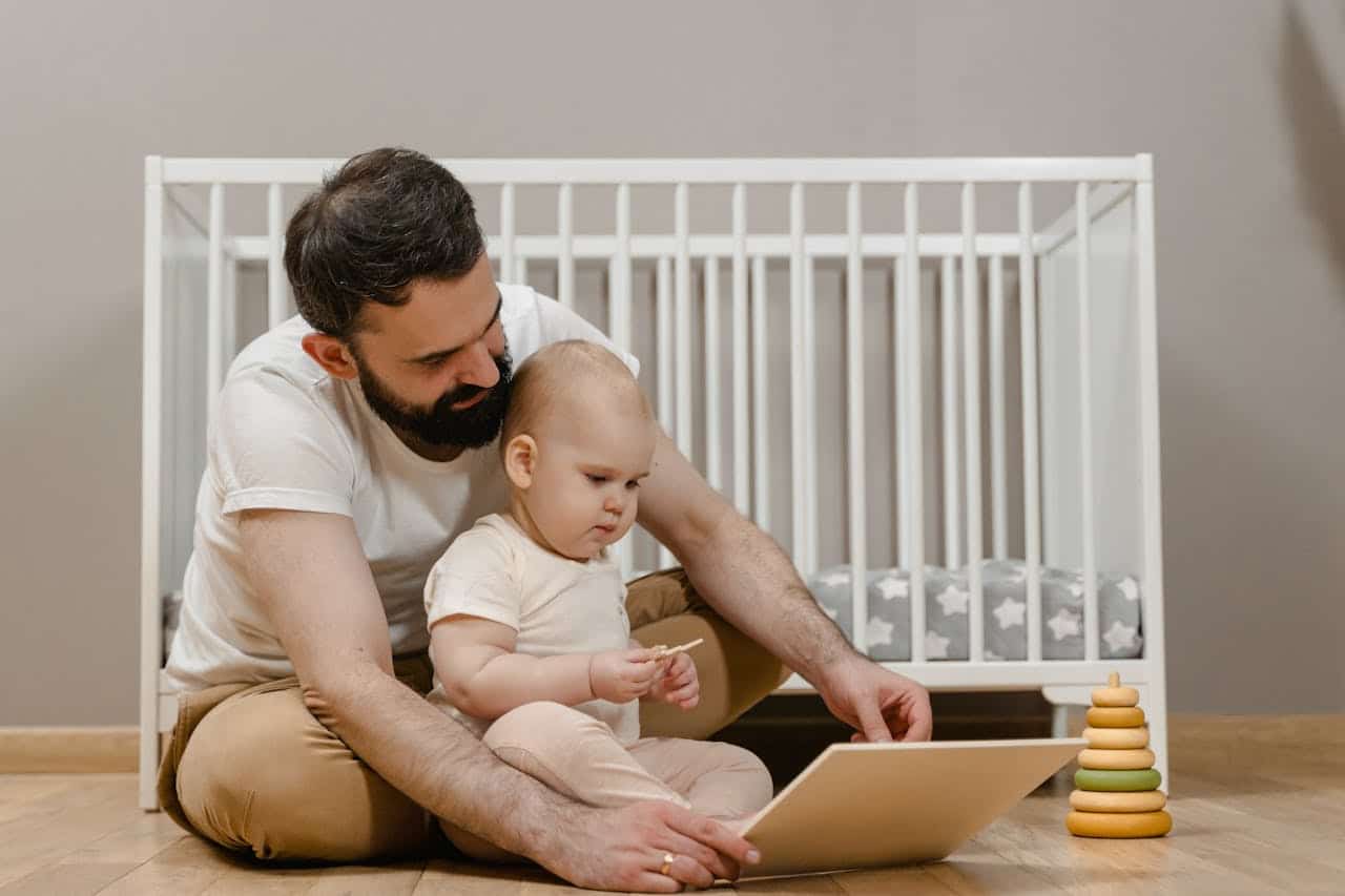 Father and baby engaging with a tablet on the floor, with a crib and colorful stacking toy in the background, illustrating family interaction and early childhood learning.