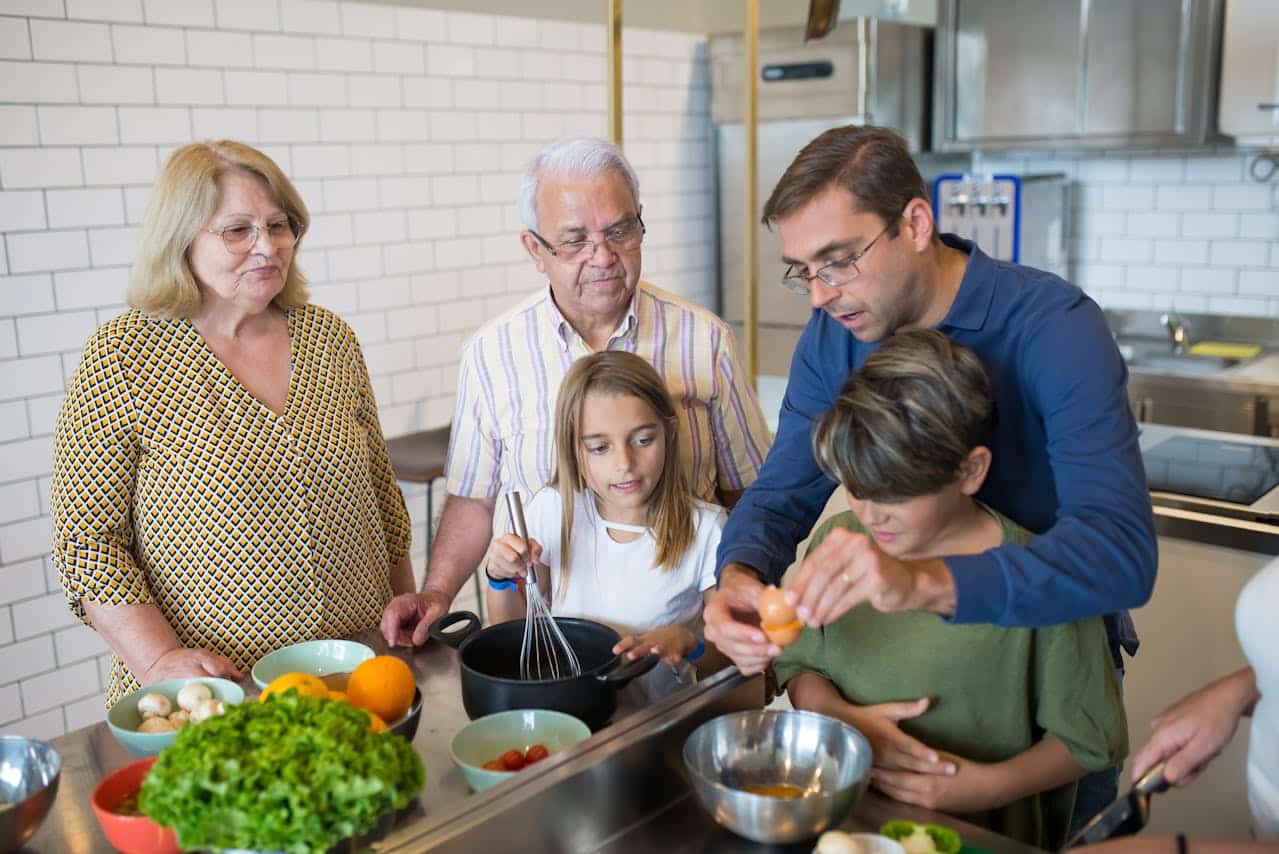 Group of four people, including two children, engaging in cooking activity in a modern kitchen, with various ingredients like lettuce, tomatoes, and oranges on the counter, emphasizing family interaction and culinary learning.