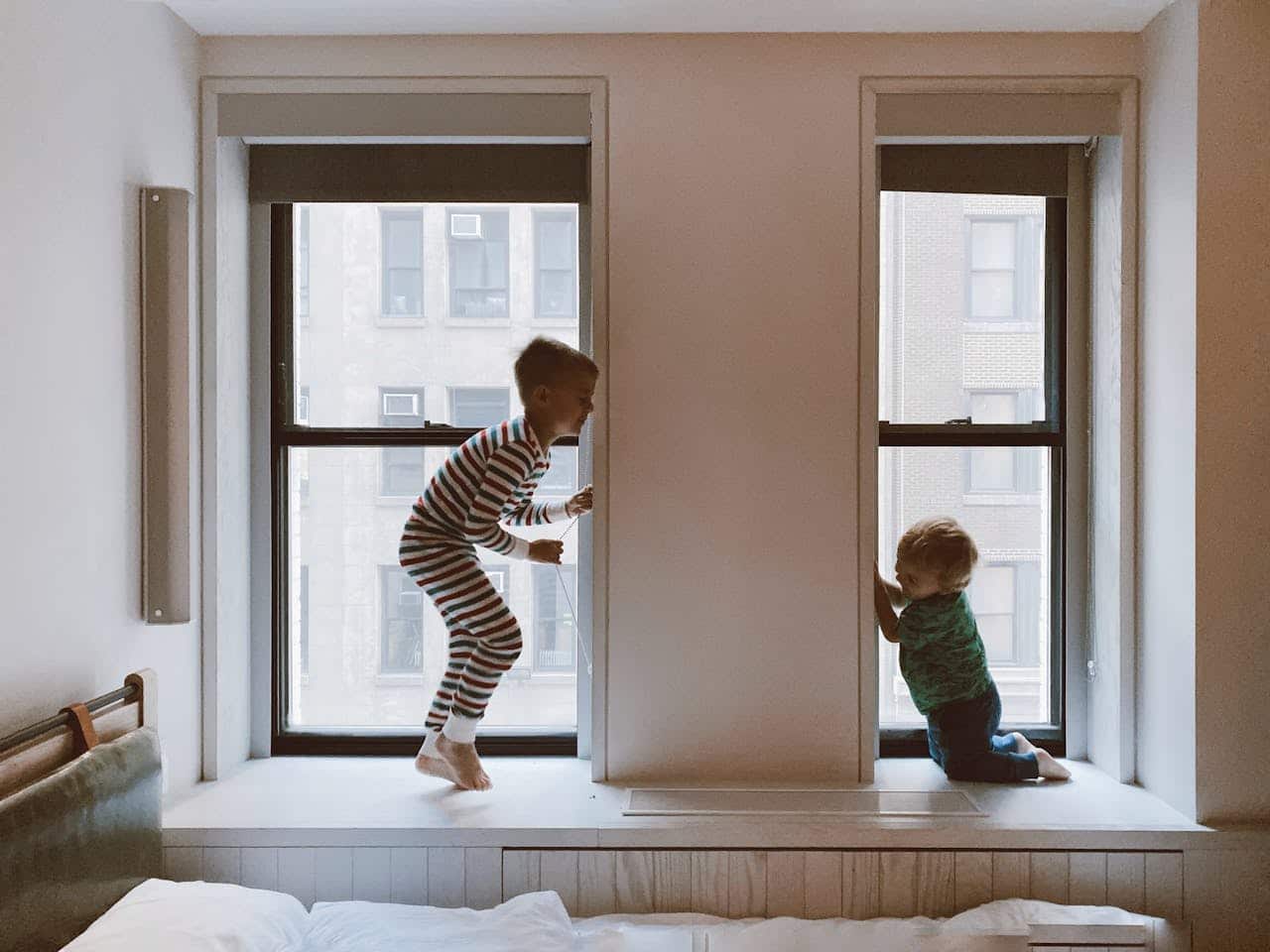 Two young boys playing in a cozy room, one jumping excitedly on a window ledge in striped pajamas, while the other kneels beside the window, both engaged in playful exploration.