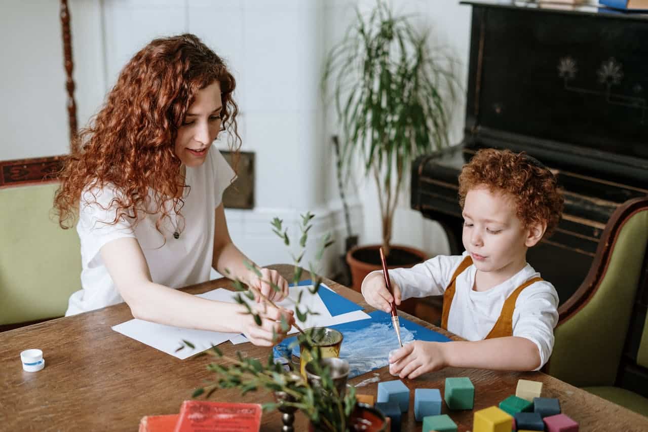 Mother and child engaging in creative activities, painting together at a wooden table, surrounded by art supplies and colorful building blocks, emphasizing family interaction and creativity.