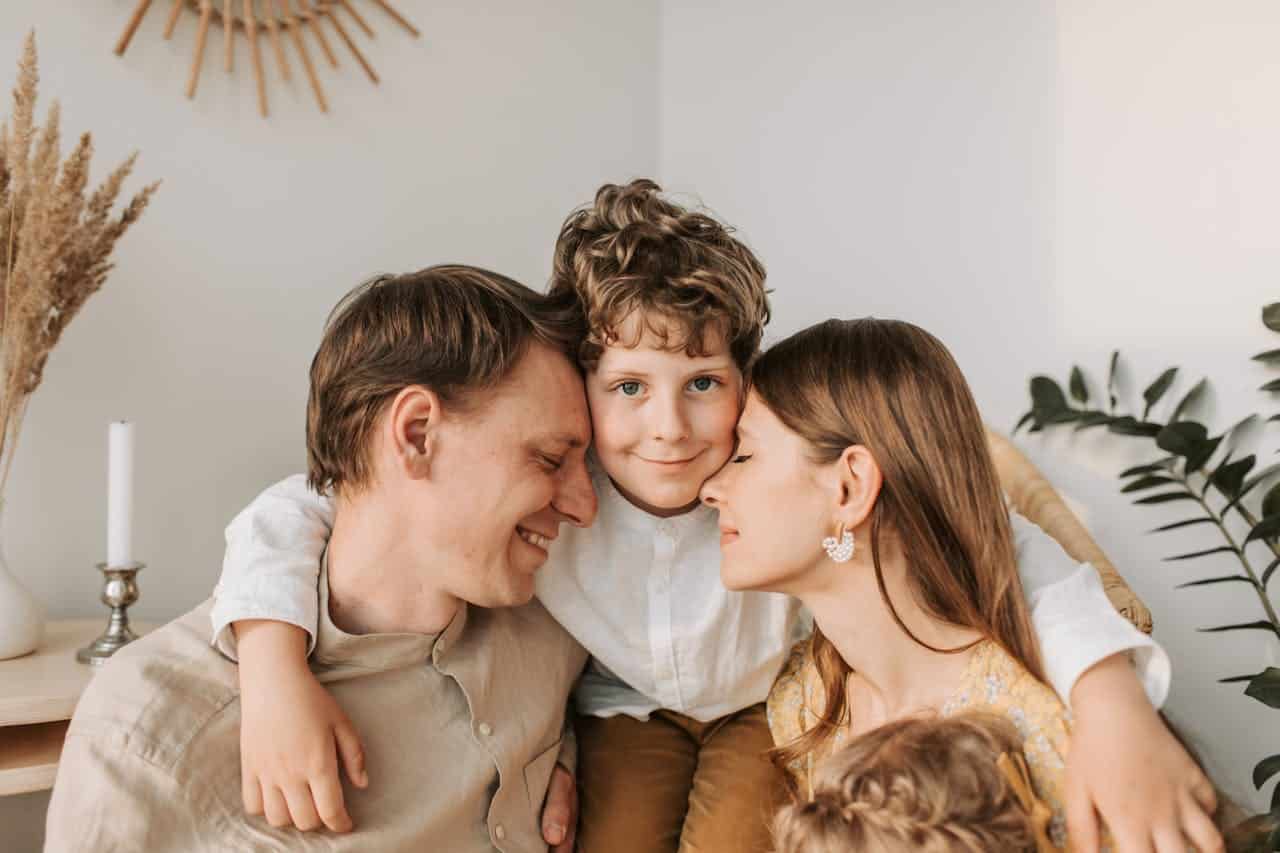 Family portrait with a smiling boy nestled between parents, showcasing affection and connection in a cozy, modern setting with plants and soft lighting.