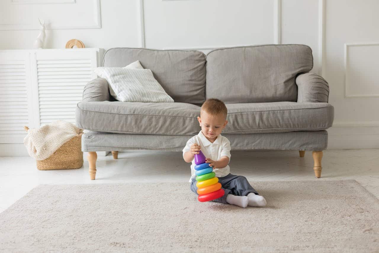 Child playing with a colorful stacking toy on a soft rug in a bright living room with a gray sofa and decorative pillows.