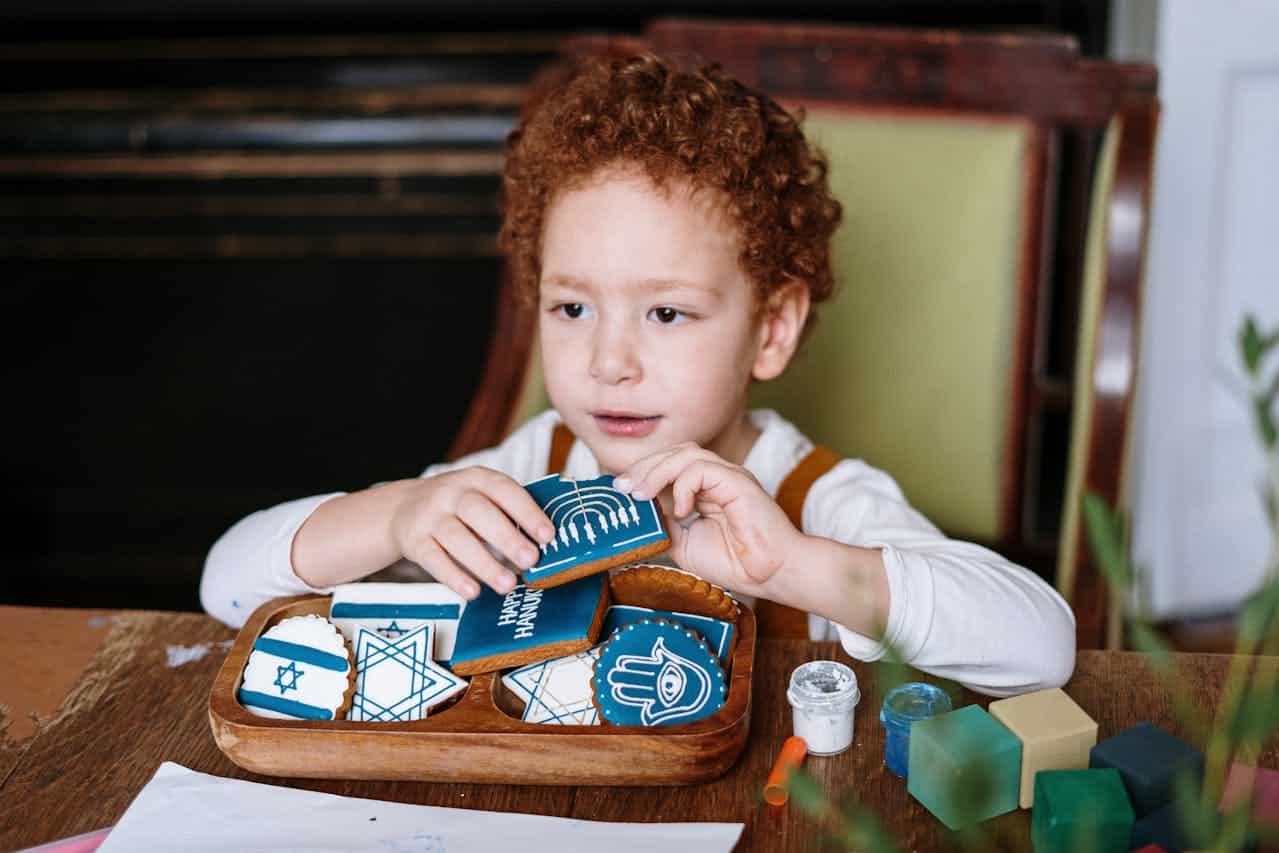 Child arranging blue and white Hanukkah-themed cookies on a wooden tray, with art supplies and colorful blocks on a table, reflecting festive creativity and celebration.
