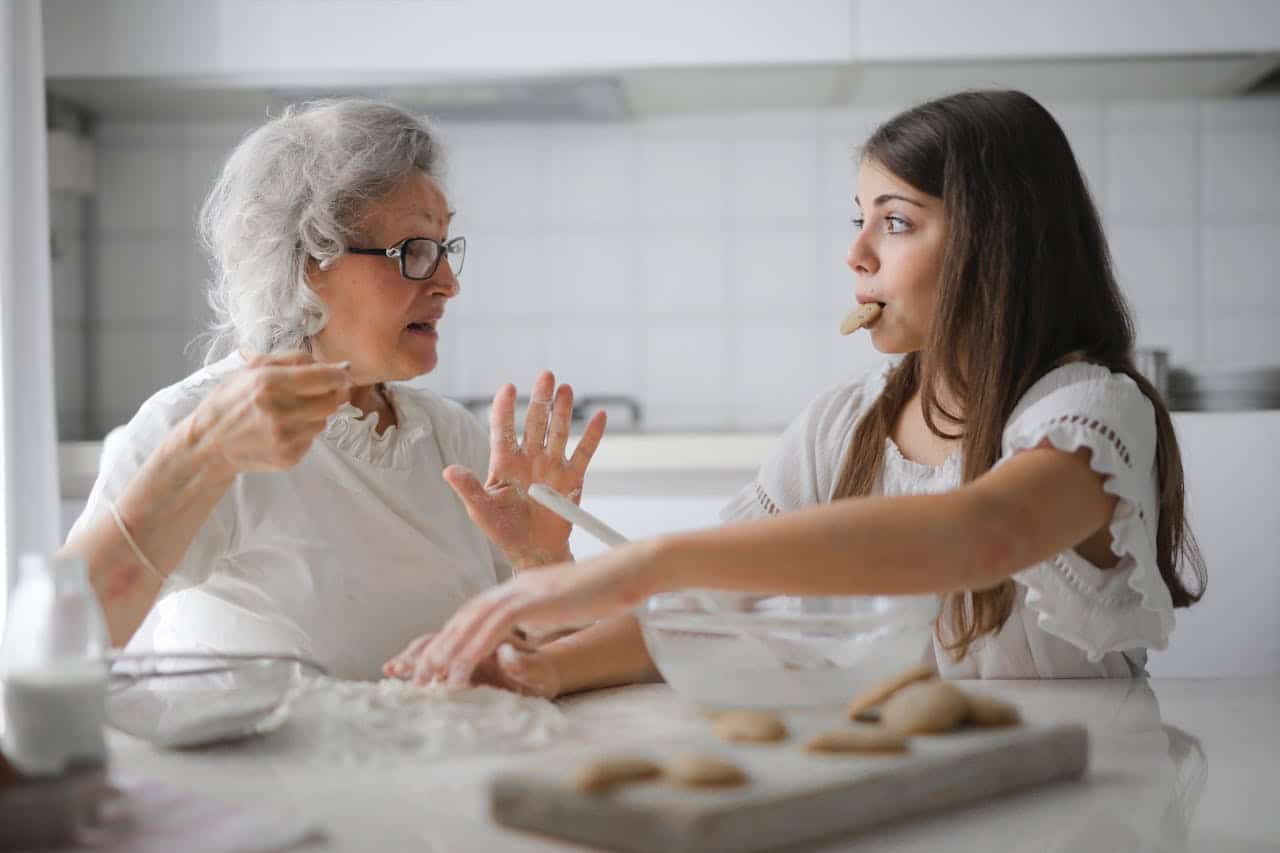 Two women in a kitchen, one elderly with gray hair and glasses gesturing while the younger woman, with long hair and a cookie in her mouth, listens attentively, surrounded by baking ingredients and cookie dough on a table.