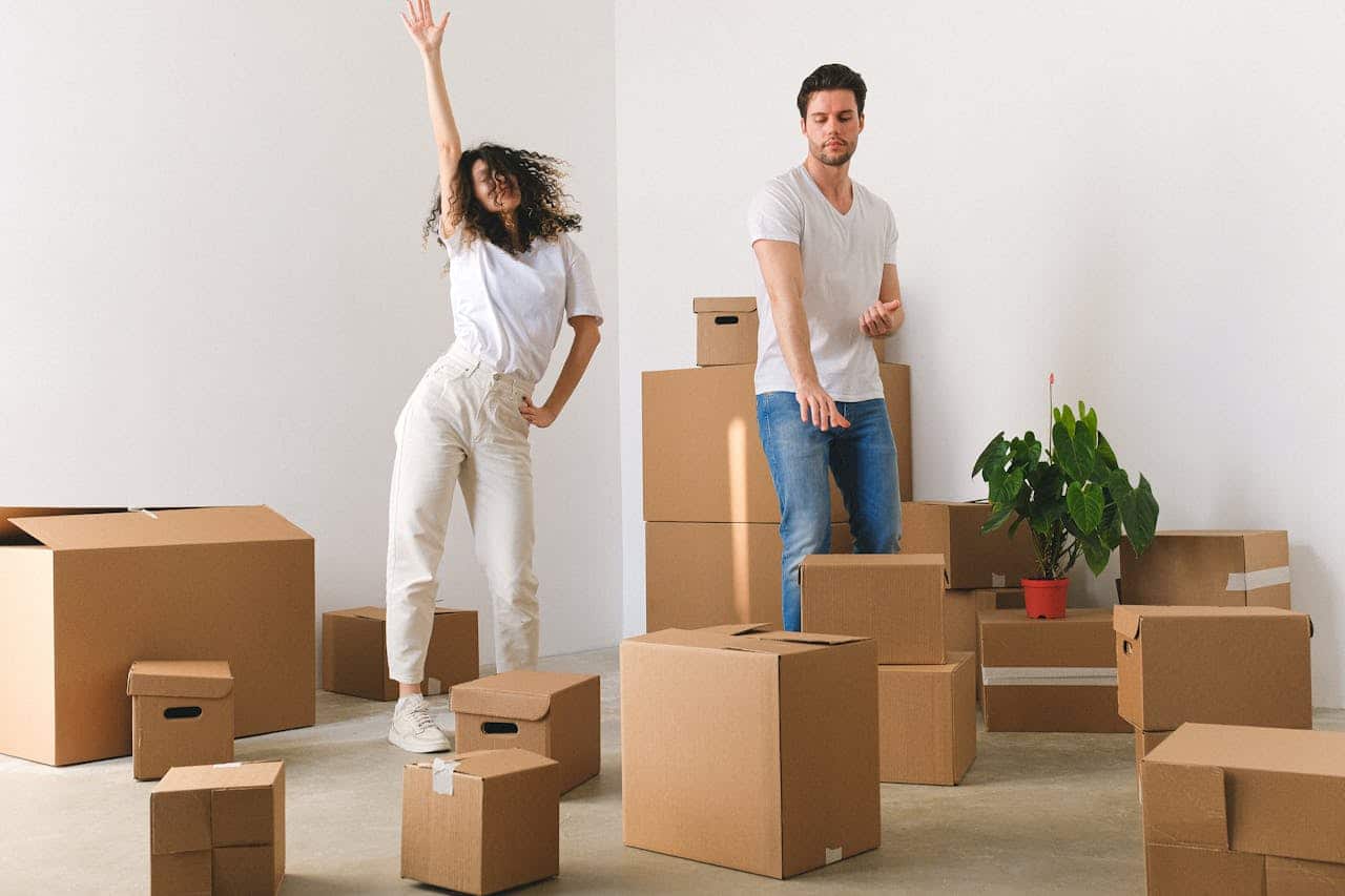 Young couple celebrating in a new home surrounded by moving boxes and a potted plant, illustrating the excitement of homeownership without traditional mortgage processes.