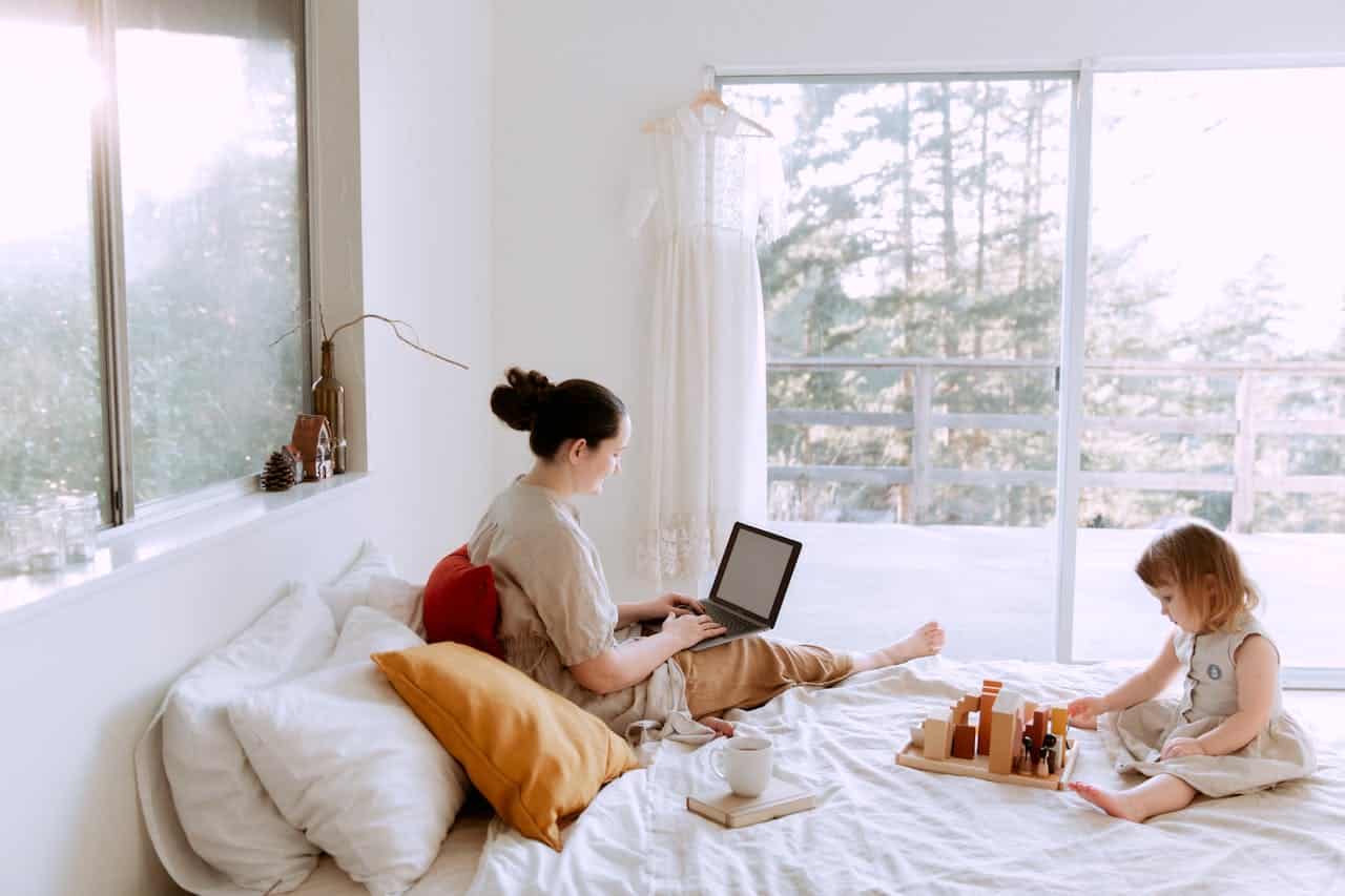 Woman working on laptop in cozy bedroom with child playing nearby, emphasizing a relaxed home environment suitable for families considering alternative mortgage options.