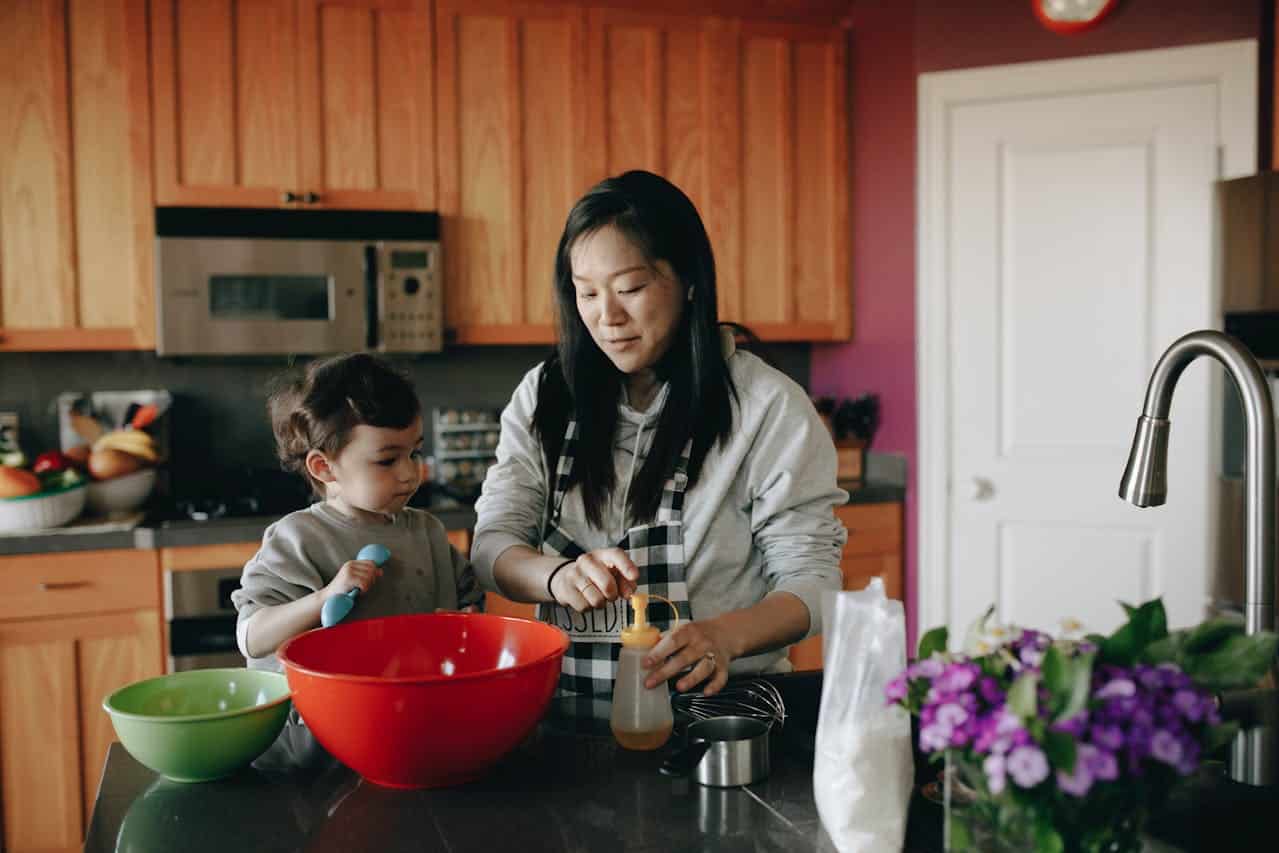 Woman and child cooking in a kitchen, using a red mixing bowl, green bowl, and measuring tools, emphasizing family engagement in food preparation.