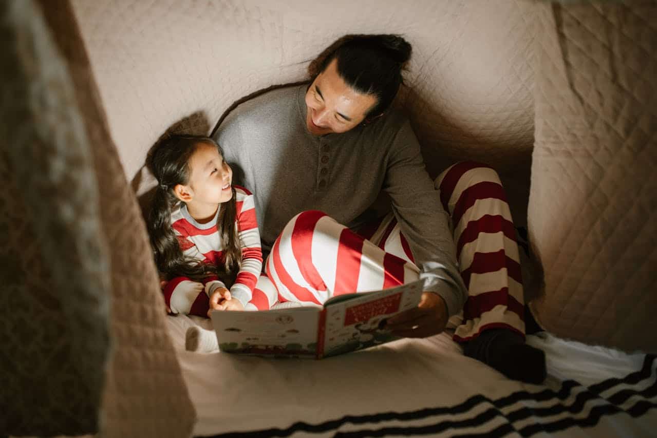 Father and daughter in cozy pajamas reading a book together in a blanket fort, smiling and enjoying quality time.