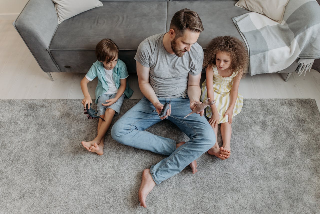 Father playing with children on carpet, holding toy figures, in a cozy living room setting.