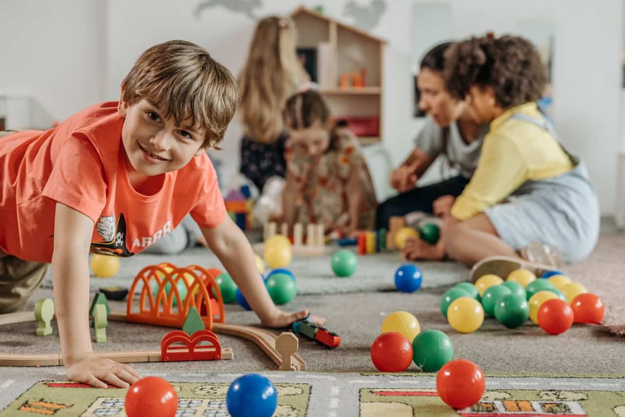 Child playing with colorful toys and building blocks in a bright, engaging playroom with other children and adults interacting nearby.