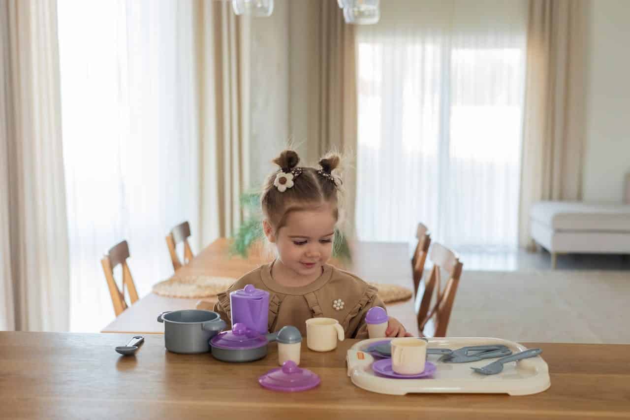 Child playing with toy kitchen set, including pots and cups, in a bright, airy dining area.