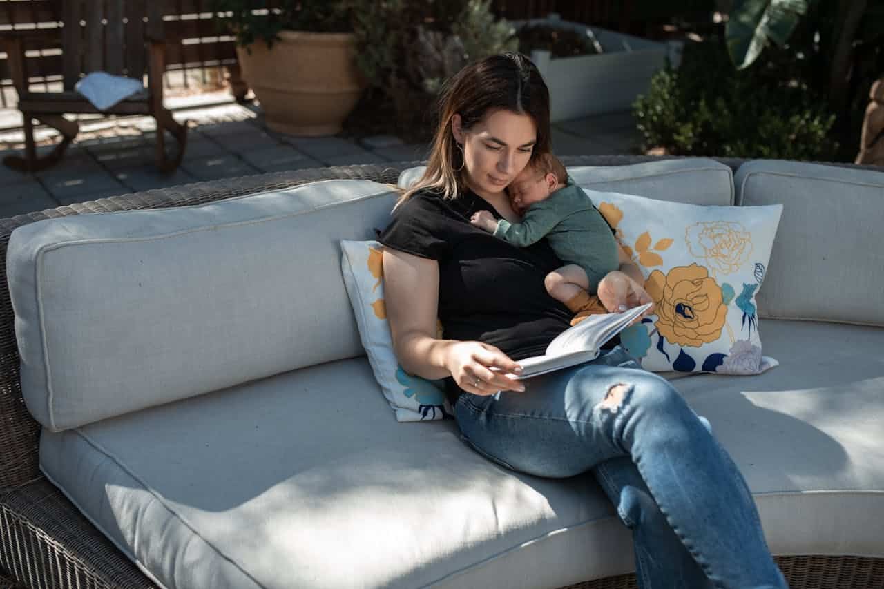 Woman reading a book on a couch while holding a sleeping baby, surrounded by decorative pillows in a serene outdoor setting.