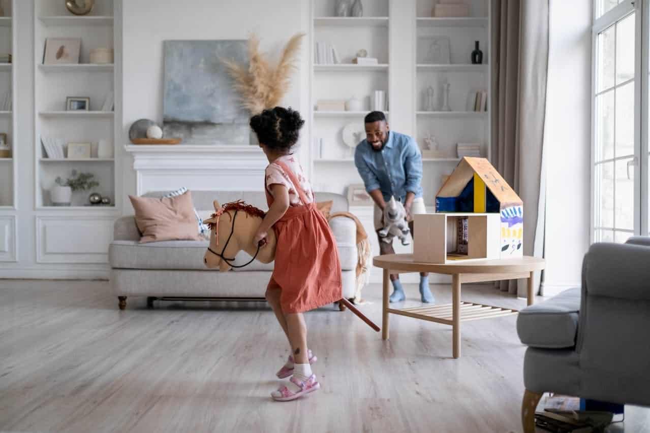 Child playing with a toy horse in a bright living room, while an adult smiles and carries a toy, illustrating a family-friendly environment ideal for homeownership discussions.