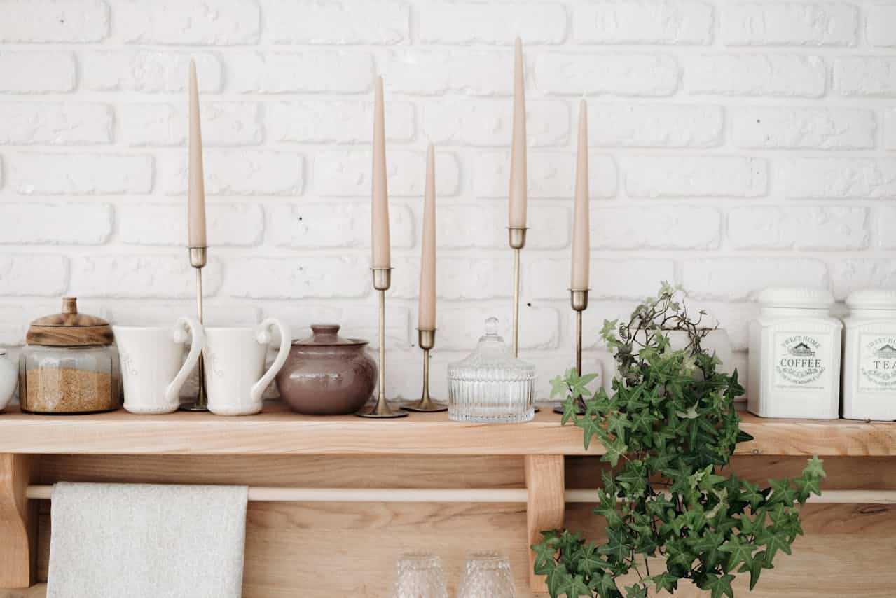 Decorative kitchen shelf with candles, ceramic mugs, a sugar jar, a teapot, glass containers, and potted ivy against a white brick wall, enhancing homeownership ambiance.