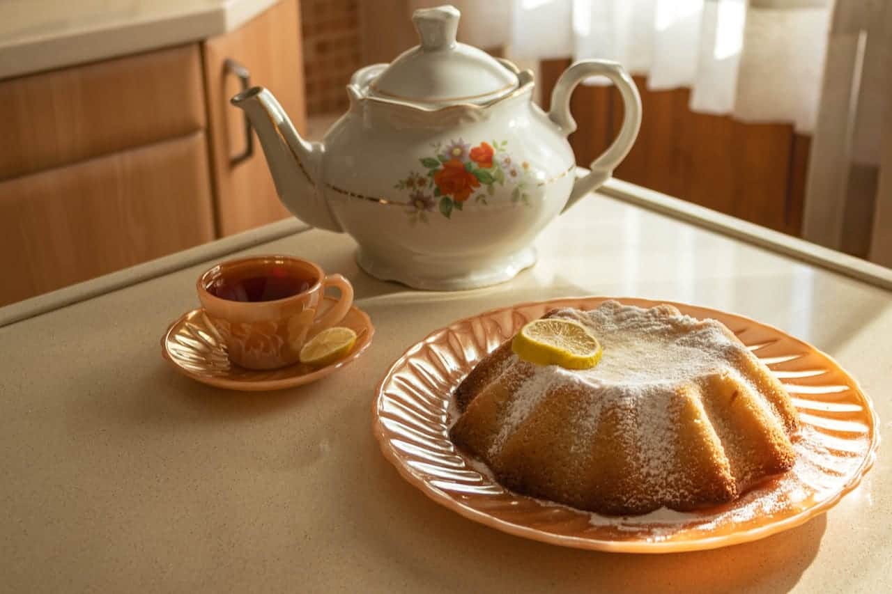 Tea set with a floral teapot, a cup of tea on a pink saucer, and a lemon-topped bundt cake dusted with powdered sugar on a matching plate, set on a kitchen counter.