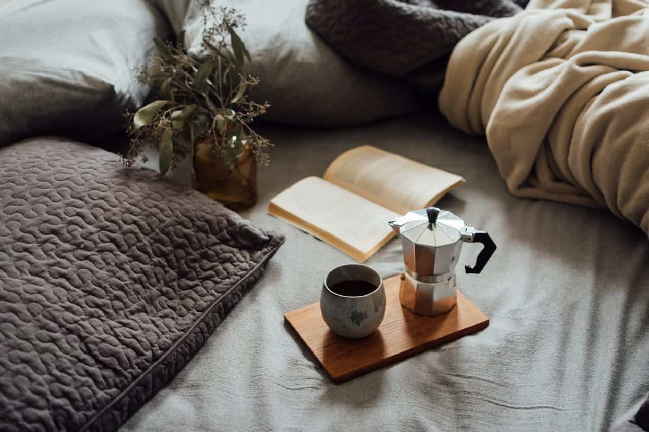 Coffee pot and cup on wooden tray, open book nearby, cozy bedding with pillows, and dried flowers in a vase, creating a warm, inviting atmosphere for relaxation and reading.