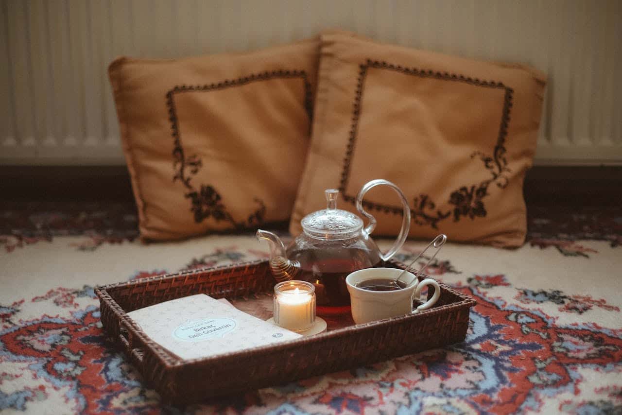 Tea set on a woven tray with a glass teapot, a cup of tea, a lit candle, and a book, surrounded by decorative pillows on a patterned rug.