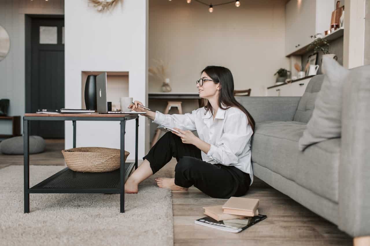 Woman sitting on the floor beside a desk with a laptop, taking notes and enjoying a cup of coffee in a cozy home office setting, reflecting the theme of owner financing and homeownership.