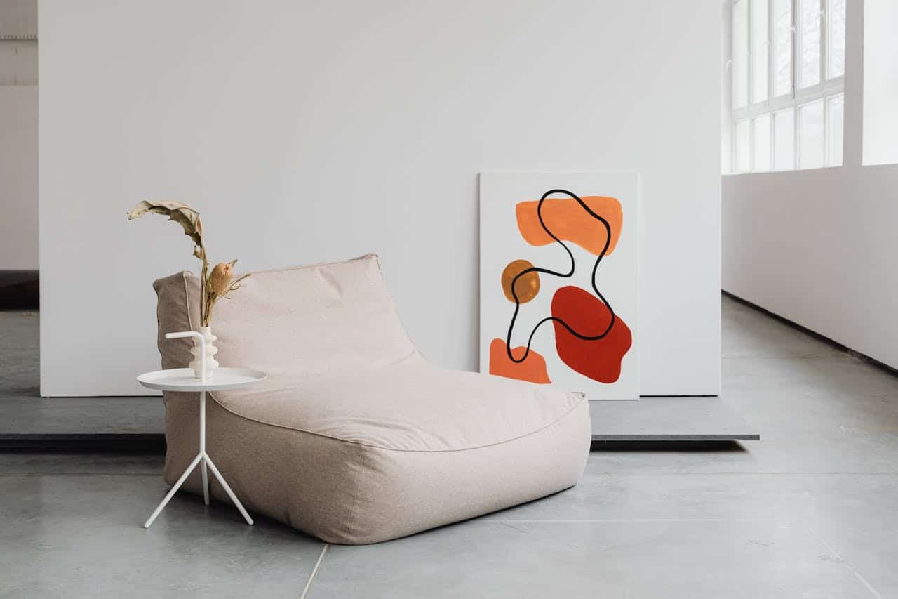 Cozy, light-colored bean bag chair beside a minimalist white side table with a decorative vase, featuring abstract art on the wall, creating a modern home interior setting.